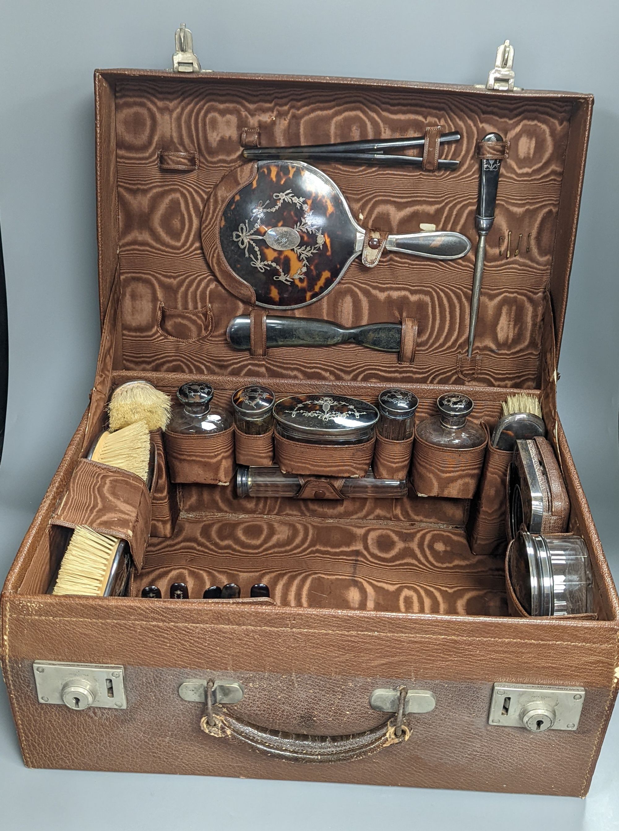 A George V leather travelling toilet case, containing twenty one matched, mainly silver and tortoiseshell pique items including toilet jars, brushes, mirror and pocket watch holder, mostly Levi & Salaman, Birmingham, 191